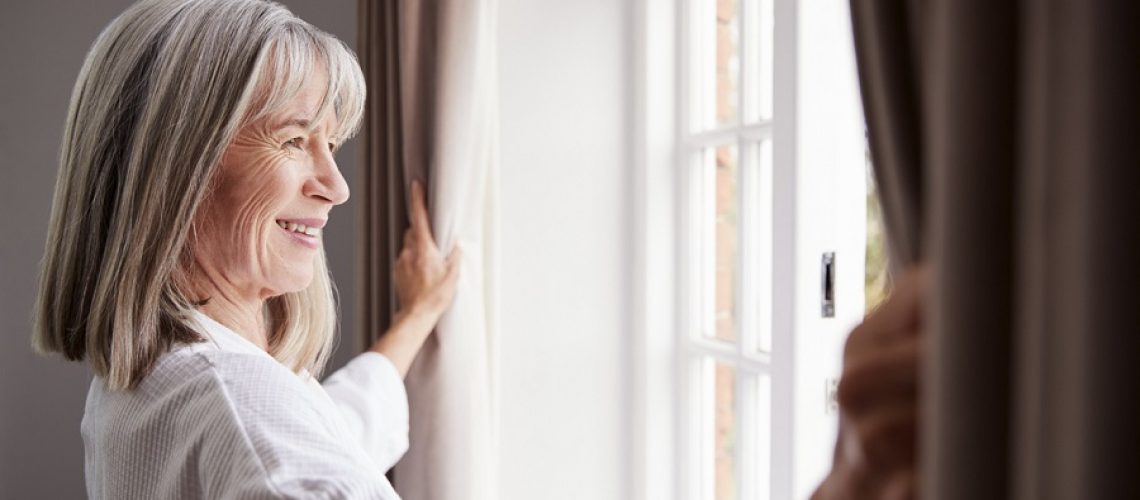 Senior Woman Opening Bedroom Curtains And Looking Out Of Window